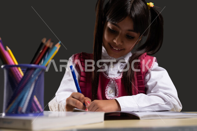 حب العلم والمعرفة، دور المدارس في التشجيع على الابتكار والاستكشاف، بورتريه مقرب لفتاة عربية سعودية خليجية ترتدي  المريول المدرسي تجلس على طاولة الدراسة وتكتب على الدفتر، أداء الواجبات المدرسية بجد واجتهاد، خلفية سوداء.