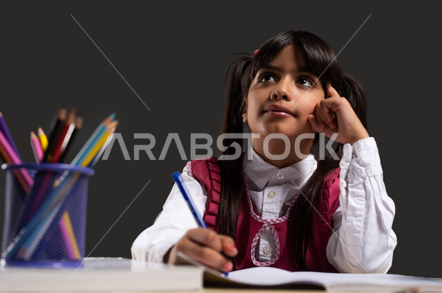تعزيز الابتكار والابداع في المدارس، ايماءات التفكير  بحل المسألة، بورتريه مقرب لطالبة عربية سعودية خليجية ترتدي المريول المدرسي تجلس على طاولة الدراسة وتنظر الى شيء ما، النمو العقلي السليم لطلاب المدارس، خلفية سوداء.