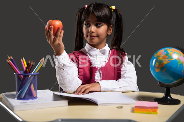 تناول الطعام الصحي للنمو العقلي والجسدي السليم، السعي لتحقيق الطموحات والأهداف العلمية، بورتريه مقرب لفتاة عربية سعودية خليجية ترتدي المريول المدرسي تجلس على طاولة الدراسة وتحمل بيدها تفاحة طازجة، خلفية سوداء.