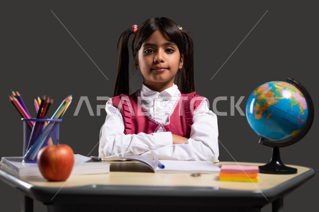ايماءات الاهتمام والجدية في تحقيق الأهداف العلمية ، التعليم في المملكة العربية السعودية، العودة الى المدارس، بورتريه مقرب لفتاة عربية سعودية خليجية ترتدي الزي المدرسي وتجلس تجلس على طاولة الدراسة مكتفة اليدين، خلفية سوداء.