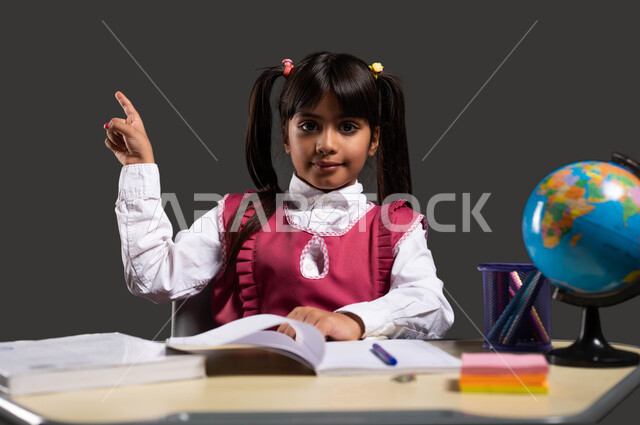 العودة الى المدارس، بورتريه مقرب لتلميذة عربية سعودية خليجية ترتدي الزي المدرسي تجلس على طاولة الدراسة وتشير بيدها الى شيء ما، تطوير المناهج والمواد التعليمية في السعوديه، رؤية مستقبلية سعودية 2030، خلفية سوداء.