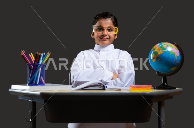 The role of schools in enhancing students’ abilities, developing curricula and educational materials in Saudi Arabia, love of learning and teaching, close-up portrait of a smiling Saudi-Gulf Arab student wearing traditional clothing and glasses sitting at the study table and pointing his finger at something, black background