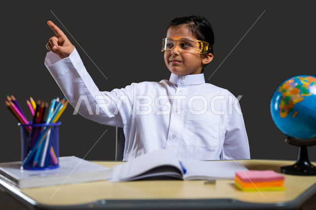 The role of schools in enhancing students’ abilities, developing curricula and educational materials in Saudi Arabia, love of learning and teaching, close-up portrait of a smiling Saudi-Gulf Arab student wearing traditional clothing and glasses sitting at the study table and pointing his finger at something, black background