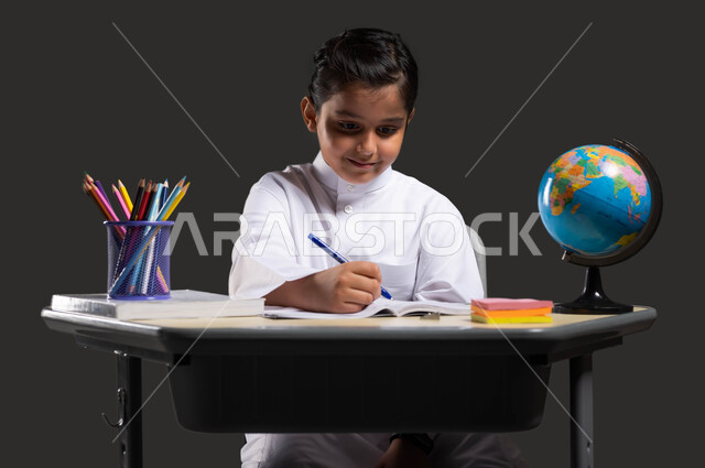 The role of schools in developing student skills, gestures of integration and concentration by reviewing daily lessons, curriculum development in Saudi Arabia, love of learning and teaching, close-up portrait of a smiling Saudi Arabian Gulf student wearing traditional clothing sitting at the study table and holding a pen in his hand, black background