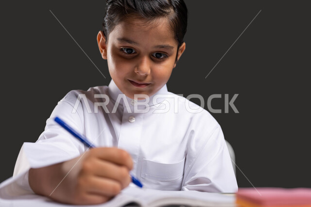 ايماءات الاندماج والتركيز بمراجعة الدروس اليومية، حب التعلم والتعليم، دور المدارس في بناء القيم والأخلاق الحسنه للطلاب، بورتريه مقرب لطالب عربي سعودي خليجي مبتسم يرتدي الزي التقليدي يجلس على طاولة الدراسة ويمسك بيده القلم، خلفية سوداء