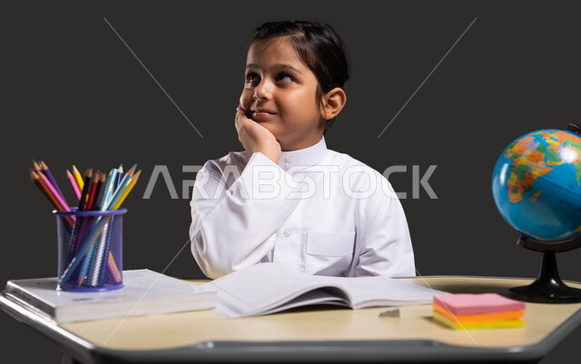 Gestures of thinking and contemplation, developing curricula and educational materials in Saudi Arabia, the importance of education in developing Saudi society, the role of schools in raising generations capable of taking responsibility, a portrait of a smiling Saudi Arabian Gulf student wearing traditional clothing and sitting at the study table, black background