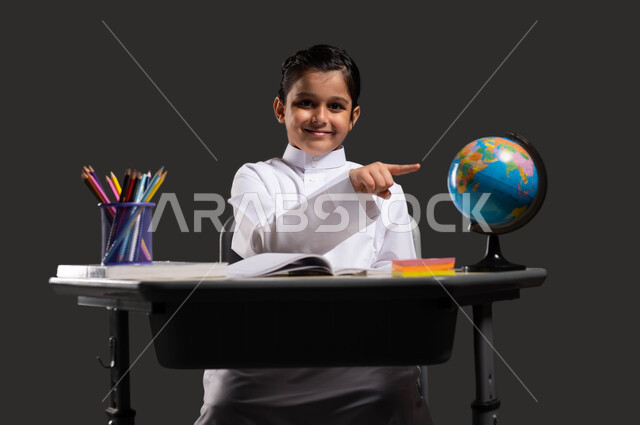 Developing curricula and educational materials in Saudi Arabia, the importance of education in developing Saudi society, the role of schools in raising generations capable of taking responsibility, a portrait of a smiling Saudi Arabian Gulf student wearing traditional clothing and pointing to a globe, black background.