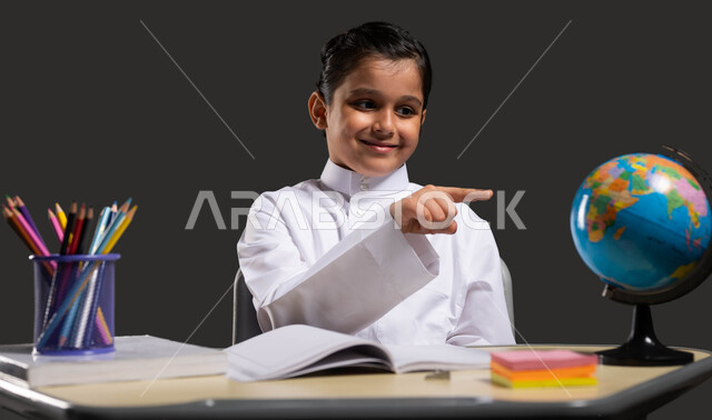 The importance of education in developing Saudi society, the role of schools in raising generations capable of taking responsibility, a close-up portrait of a smiling Saudi-Gulf Arab student wearing traditional clothing and pointing to the globe, developing curricula and educational materials in Saudi Arabia, black background