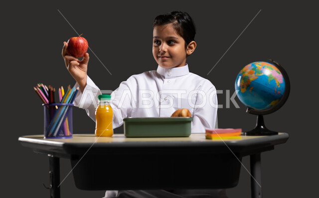 The importance of education in building a bright future for Saudi society, the role of schools in raising generations capable of taking responsibility and developing society, a close-up portrait of a smiling Saudi-Gulf Arab student wearing traditional clothing and holding an apple in his hand, developing curricula and educational materials in Saudi Arabia, black background