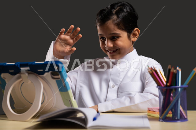 Gestures of pleasure in participating with the teacher via live broadcast, interactive learning via the Internet, modern and developed educational curricula in the Kingdom, a close-up portrait of a Saudi Gulf Arab boy wearing traditional Saudi clothing, using a tablet to follow the explanation of school subjects, black background.