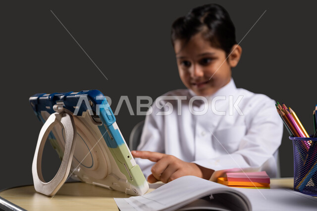 ايماءات الاندماج والتركيز، تفاعل الطلاب مع المحتوى التعليمي عبر الإنترنت، المناهج التعليمية الحديثة والمطورة بالمملكة ، بورتريه مقرب لولد عربي سعودي خليجي يرتدي الزي السعودي التقليدي  يقوم باستخدام الجهاز اللوحي، خلفية سوداء