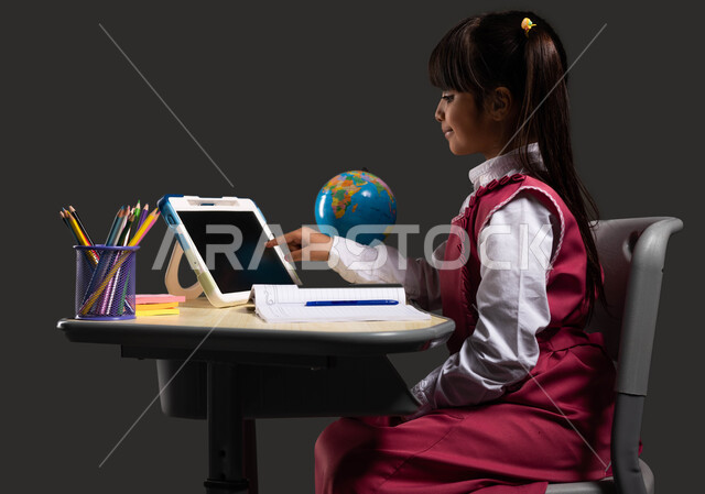 Interactive online learning using a tablet, the role of technology in changing traditional education methods, a portrait from the side of a Saudi Arabian Gulf student wearing a school uniform and sitting at the study table, black background.