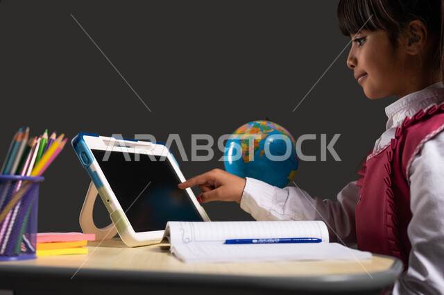 Active online learning using a tablet, modern and developed educational courses and curricula in the Kingdom, a close-up portrait from the side of a Saudi Arabian Gulf student wearing a school uniform, sitting at the study table and pointing to the tablet, black background.