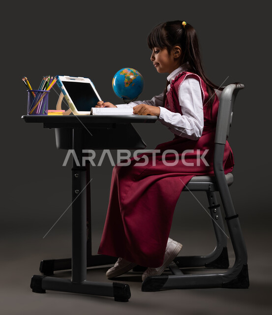 المقررات والمناهج التعليمية الحديثة والمطورة  في السعودية، التعلم النشط عبر الإنترنت، بورتريه من الجانب لطالبة عربية سعودية خليجية ترتدي الزي المدرسي وتجلس على طاولة الدراسة، الدراسة المدرسية الفعالة عبر الجهاز اللوحي، خلفية سوداء.