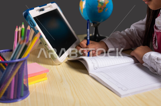 Reviewing daily lessons online, the development of science and education in the Kingdom of Saudi Arabia, a portrait from the side of a Saudi Gulf Arab student wearing a school uniform and sitting at the study table in front of the tablet, black background.