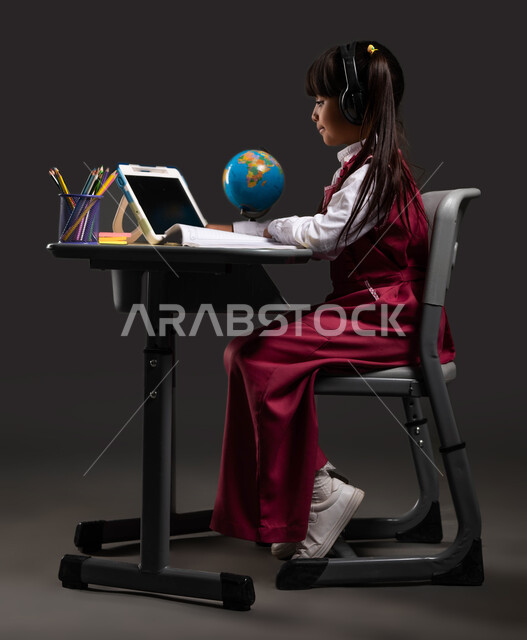 Doing schoolwork online, the development of education in the Kingdom of Saudi Arabia, a side portrait of a Saudi Arabian Gulf student wearing a school uniform, listening to lessons via advanced headphones, sitting at the study table in front of the tablet, black background.