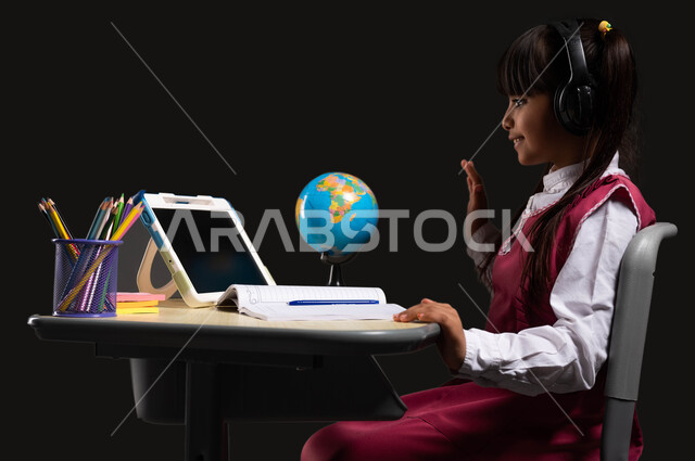 التفاعل والمشاركه في الحصص اثناء البث المباشر للدرس، مفهوم التعلم عبر الانترنت، بورتريه لتلميذة عربية سعودية خليجية ترتدي اليونيفورم المدرسي تضع سماعات الرأس الصوتية وتجلس على طاولة الدراسة أمام التابلت،  خلفية سوداء.