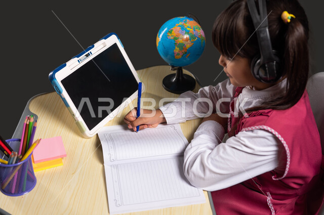 استخدام التطبيقات الرقمية في التعلم عن بعد، بورتريه مقرب لفتاة عربية سعودية خليجية ترتدي اليونيفورم المدرسي تضع سماعات الرأس الصوتية وتجلس على طاولة الدراسة أمام التابلت، مفهوم التعلم عبر الانترنت، تطور التعليم  في المملكة العربية السعودية، خلفية سوداء.