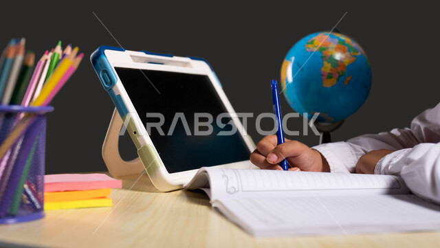 استخدام الأجهزة الذكية في التعلم عن بعد، بورتريه مقرب لتلميذة عربية سعودية خليجية ترتدي اليونيفورم المدرسي وتجلس على طاولة الدراسة أمام التابلت، مفهوم التعلم عبر الانترنت، تطور التعليم في المملكة العربية السعودية، خلفية سوداء.