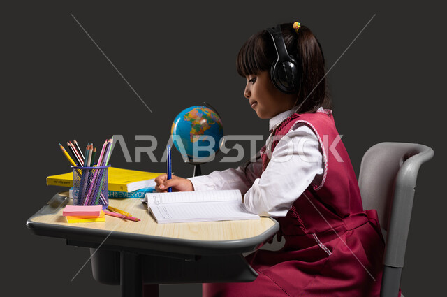 الحرص على التفوق والنجاح، مفهوم التعليم الصوتي السماعي، استخدام القرطاسية والادوات المدرسية، التعلم وسماع الدروس صوتيا، بورتريه مقرب من الجانب لطالبة عربية خليجية سعودية ترتدي الزي المدرسي تضع سماعات الرأس وتجلس على الطاولة وتقوم بحل واجباتها، خلفية سوداء.