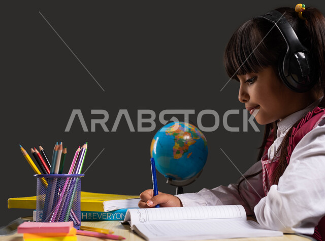 Eagerness for excellence and success, the concept of audio-audio education, using stationery and school tools, learning and listening to lessons audio, a close-up portrait from the side of a Saudi Arabian Gulf student wearing a school uniform, wearing headphones, sitting at the table and doing her homework, black background.