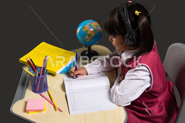 مفهوم التعليم الصوتي السماعي، استخدام القرطاسية والادوات المدرسية، التعلم وسماع الدروس صوتيا، بورتريه من الجانب لطالبة عربية خليجية سعودية ترتدي الزي المدرسي تضع سماعات الرأس وتجلس على الطاولة تقوم بحل واجباتها، خلفية سوداء.