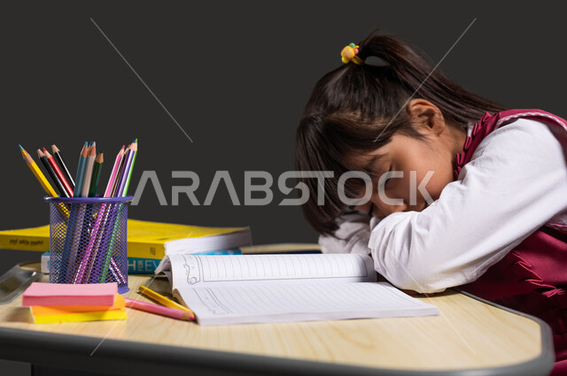 Returning to school in Saudi Arabia, feeling lazy and lethargic to complete school tasks and duties, the need to sleep and take a rest, a close-up portrait of a Saudi Arabian Gulf girl wearing a school dress sleeping at the study table, sleepy gestures and a feeling of fatigue, black background.