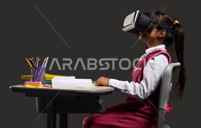 مفهوم التعليم المنزلي، متابعة شرح الدرس عبر الفي ار، أساليب تعليم متطورة باستخدام التقنيات الحديثة، بورتريه لفتاة عربية خليجية سعودية ترتدي الثوب المدرسي تجلس أمام الطاولة الدراسية وتضع نظارة الواقع الإفتراضي ثلاثية الأبعاد، خلفية سوداء.