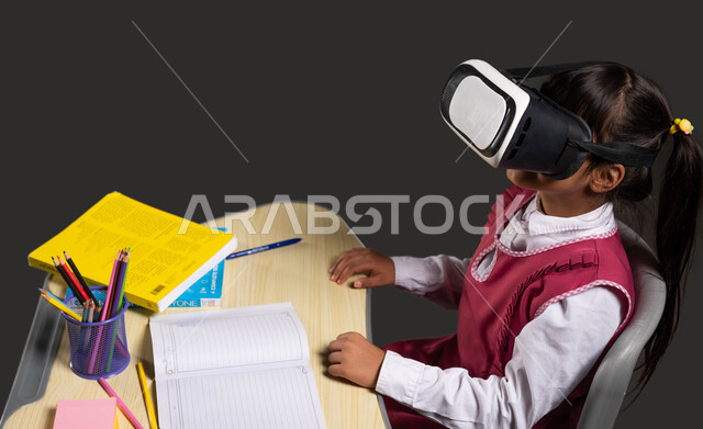 The concept of home education, following the explanation of the lesson via VR, advanced teaching methods using modern technologies, a portrait of a Saudi Gulf Arab girl wearing a school dress sitting in front of the study table and wearing 3D virtual reality glasses, a black background.