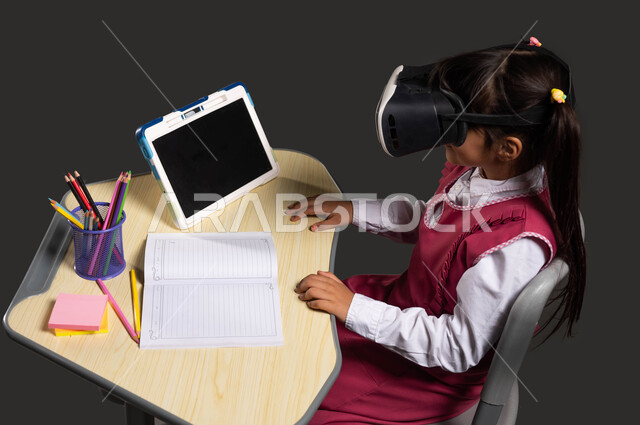استخدام التقنيات الحديثة والمتطورة ومحاكاة العالم الخيالي في التعليم، بورتريه لفتاة عربية خليجية سعودية ترتدي الثوب المدرسي تجلس أمام الطاولة الدراسية والتابلت وتضع نظارة الواقع الإفتراضي ثلاثية الأبعاد، ايماءات الاندماج والتركيز ومشاهدة شيء ما، خلفية سوداء.