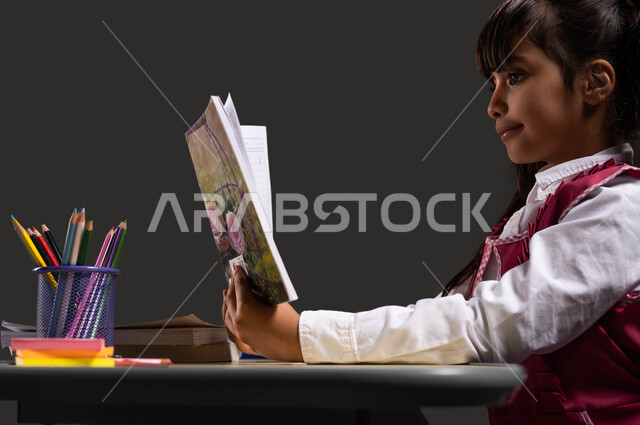Diligence, striving for glory, perseverance, and eagerness to succeed, gestures indicating focus, close-up portrait from the side of a Saudi Arabian Gulf student wearing a school uniform, sitting at the table studying for tests and preparing for the exam, black background.
