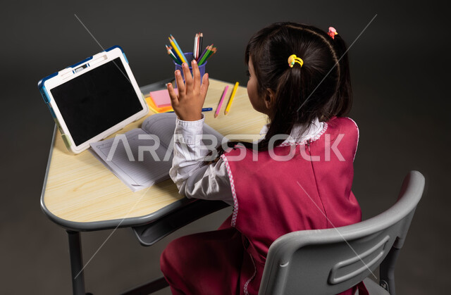 The concept of online learning, modern and advanced educational methods and curricula in Saudi Arabia, following lessons through distance education platforms, a portrait of a Saudi Arabian Gulf student wearing a school uniform and participating via live broadcast on a tablet, a black background.
