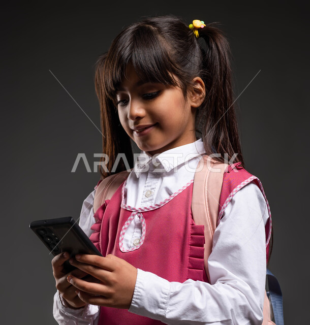 Solving and submitting assignments online, the concept of distance learning using modern technology, preparing to go to school, a portrait of a Saudi Gulf student wearing a school uniform and bag holding a mobile phone in her hand, studying through e-learning platforms, a black background.