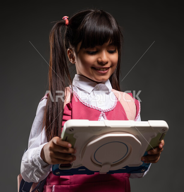 متابعة الدروس عن طريق منصات التعلم عن بعد، مفهوم التعليم المنزلي، مناهج تعليمية حديثة ومتطورة، متابعة الدروس عن طريق منصات التعلم عن بعد، بورتريه مقرب لطالبة عربية سعودية مبتسمة ترتدي الزي المدرسي والحقيبة المدرسية تحمل بيدها جهاز التابلت التقني، خلفية سوداء.