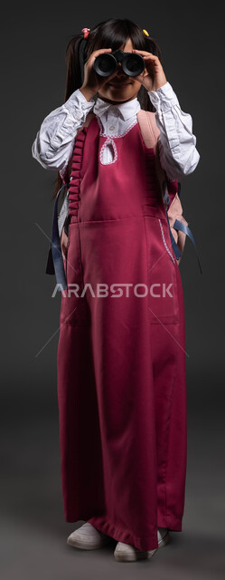 صورة الجسم بالطول الكامل، حب الاستكشاف والمغامرة، العودة إلى المدارس، جودة التعليم في السعودية، بورتريه مقرب لتلميذة عربية سعودية ترتدي الزي المدرسي والحقيبة المدرسية تضع المنظار على عينيها لتقريب الاشياء البعيدة، الاستعداد للفصل الدراسي الجديد، خلفية سوداء.