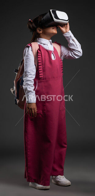 ايماءات الاندماج والتركيز، تطور أساليب التعليم باستخدام التقنيات الحديثة والمتطورة ومحاكاة العالم الخيالي، بورتريه لفتاة عربية خليجية سعودية ترتدي الثوب المدرسي والحقيبة المدرسية تضع نظارة الواقع الإفتراضي ثلاثية الأبعاد، خلفية سوداء.