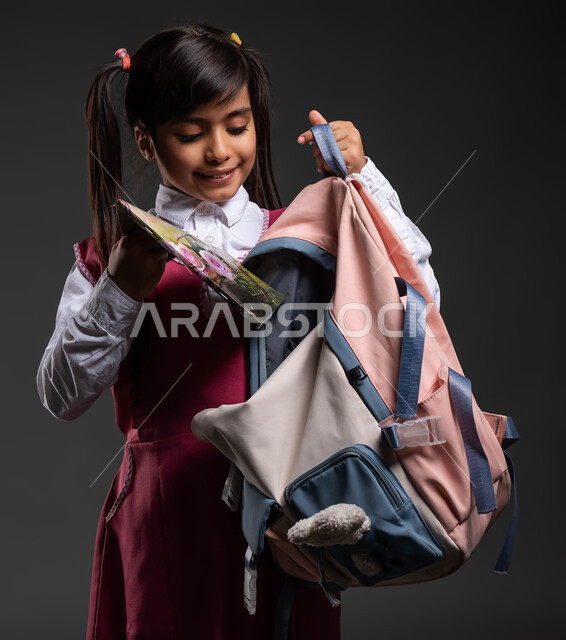 تجهيز المستلزمات المدرسية , التحضير والاستعداد للذهاب إلى المدرسة, بورتريه مقرب لفتاة عربية سعودية مبتسمة ترتدي اللباس المدرسي وتقوم بوضع الكتب الدراسية في حقيبتها, الشعور بالحماس في بداية العام الدراسي الجديد, التعليم في المملكة, خلفية رمادية.