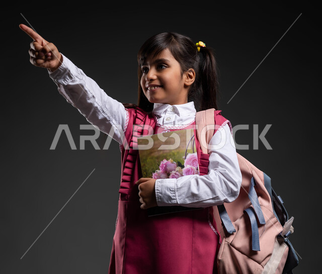 الإشارة  بإصبع السبابة إلى شيء ما, تطور وتقدم المناهج التعليمية في المملكة العربية السعودية, إيماءات الفرح بالنجاح وتحقيق الأهداف, بورتريه لفتاة عربية سعودية خليجية مبتسمة ترتدي الثوب المدرسي والحقيبة وتحمل بيدها الكتب الدراسية, خلفية سوداء.