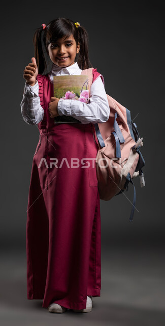 النظر إلى الكاميرا ورفع الإبهام نحو الأعلى بعلامة النجاح والتفوق, إيماءات الإعجاب بجودة التعليم في السعودية, بورتريه لفتاة سعودية مبتسمة ترتدي الزي المدرسي والحقيبة وتحمل بيدها الكتب الدراسية, بداية العام الدراسي الجديد، صورة الجسم بالطول الكامل, خلفية سوداء .