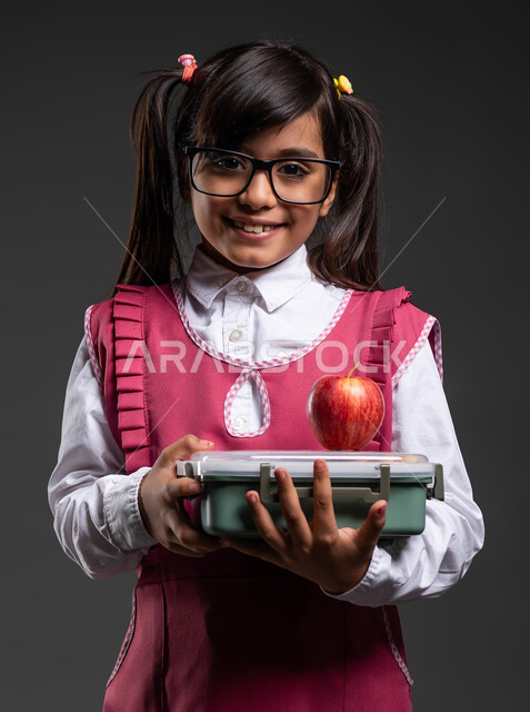 تناول وجبة فطور صحية في وقت الفسحة المدرسية, النظر إلى الكاميرا,  بورتريه مقرب لفتاة سعودية مبتسمة ترتدي اللباس المدرسي والنظارة الطبية تحمل بيدها حافظة الطعام, أكل الفواكه الطازجة للشعور بالحيوية والنشاط, العودة إلى المدارس, خلفية سوداء .