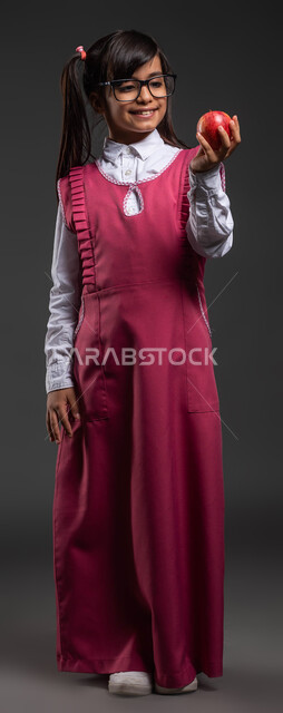 وجبة فطور صحية طازجة, تناول الطعام الصحي في وقت الفسحة, بورتريه لتلميذة عربية خليجية سعودية مبتسمة ترتدي اللباس المدرسي والنظارة الطبية تحمل بيدها تفاحة حمراء, مفهوم العناية والاهتمام بالصحة, صورة الجسم بالطول الكامل, خلفية سوداء .