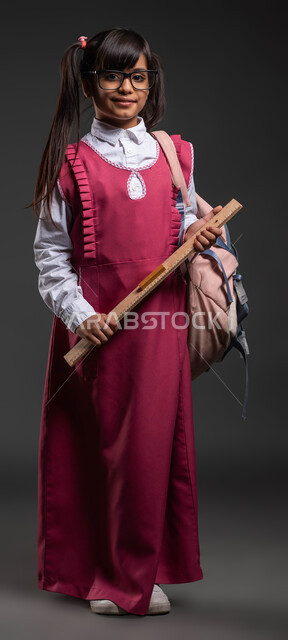 الذكاء والفطنة في الرياضيات , قرطاسية ومستلزمات مدرسية ,بورتريه لطالبة عربية خليجية سعودية مبتمسة ترتدي الزي المدرسي والنظارة الطبية تحمل الحقيبة المدرسية ومسطرة خشبية مرقمة ,الأناقة والاهتمام بالمظهر الخارجي , صورة الجسم بالطول الكامل ،خلفية سوداء .