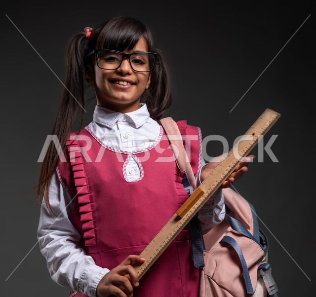فتاة مجتهدة ونشيطة , قرطاسية ومستلزمات مدرسية ,بورتريه لطالبة عربية خليجية سعودية مبتمسة ترتدي الزي المدرسي والنظارة الطبية  تحمل الحقيبة المدرسية ومسطرة خشبية مرقمة ,إيماءات النشاط والفرح بالعودة إلى المدارس ،خلفية سوداء .