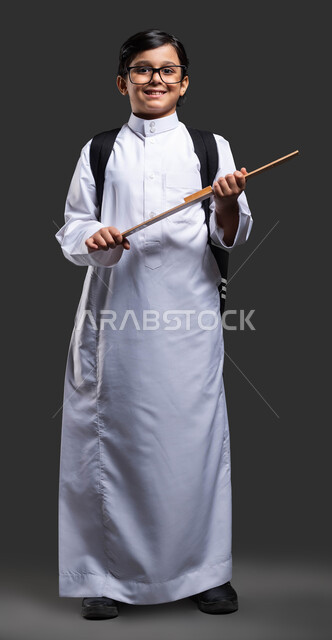 مستلزمات وقرطاسية مدرسية, بورتريه لولد عربي سعودي خليجي يرتدي الثوب المدرسي والنظارة الطبية يحمل الحقيبة المدرسية ومسطرة خشبية , بداية العام الدراسي الجديد , النظر إلى الكاميرا بإيماءات النشاط والاستعداد للدوام ,صورة الجسم بالطول الكامل , خلفية سوداء .