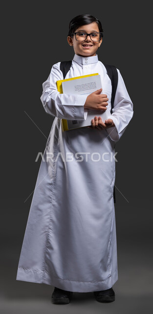 أساليب تعليمية متطورة في المملكة, مفهوم التعليم والثقافة, النظر إلى الكاميرا بإيماءات النشاط والاستعداد للدوام, بورتريه مقرب لولد عربي سعودي خليجي يرتدي الثوب المدرسي والنظارة الطبية  يحمل الحقيبة المدرسية والكتب, صورة الجسم بالطول الكامل, خلفية سوداء .