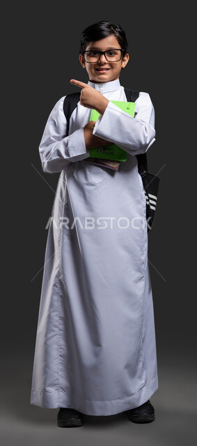 Looking at the camera with gestures of joy at success and achieving goals, pointing to something, development and progress of educational curricula in the Kingdom of Saudi Arabia, portrait of a smiling Saudi Arabian Gulf boy wearing a school uniform and carrying a school bag and textbooks, black background