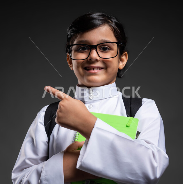 The development and progress of educational curricula in Saudi Arabia, looking at the camera with gestures of joy at success and achieving goals, portrait of a smiling Saudi Arabian Gulf boy wearing a school uniform carrying a school bag and books, pointing to something, black background