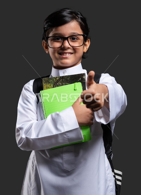 النظر إلى الكاميرا ورفع الإبهام نحو الأعلى بعلامة جودة التعليم في السعودية, تطور وتقدم المناهج التعليمية, بورتريه مقرب لولد عربي سعودي خليجي مبتسم  يرتدي الثوب المدرسي والحقيبة يحمل بيده الكتب الدراسية , النجاح والتفوق في المواد الدراسية ,  خلفية سوداء .