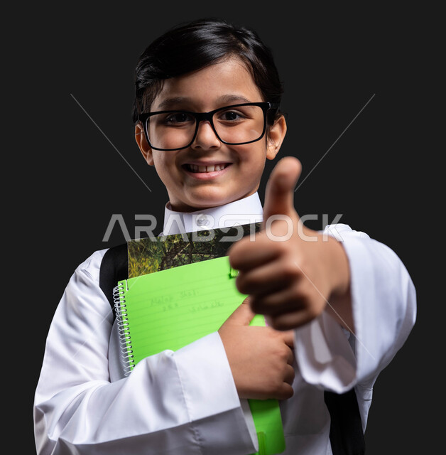 إيماءات الإعجاب بجودة التعليم في المملكة, بورتريه مقرب لولد عربي سعودي خليجي مبتسم  يرتدي الثوب المدرسي والحقيبة يحمل بيده الكتب الدراسية ,النظر إلى الكاميرا ورفع الإبهام نحو الأعلى بعلامة النجاح والتفوق , بداية العام الدراسي الجديد ,  خلفية سوداء .