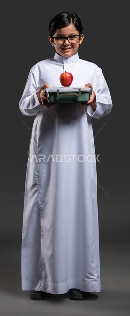 تناول الفواكه الطازجة للشعور بالحيوية والنشاط, إيماءات النظر إلى الكاميرا,تناول وجبة فطور صحية في وقت الفسحة, بورتريه لولد عربي خليجي سعودي مبتسم  يرتدي الثوب التقليدي والنظارة الطبية يحمل بيده حافظة الطعام المدرسية, صورة الجسم بالطول الكامل, خلفية سوداء .